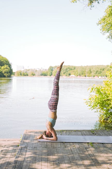 Woman in a white outfit performing a balancing yoga pose outdoors near calm water.